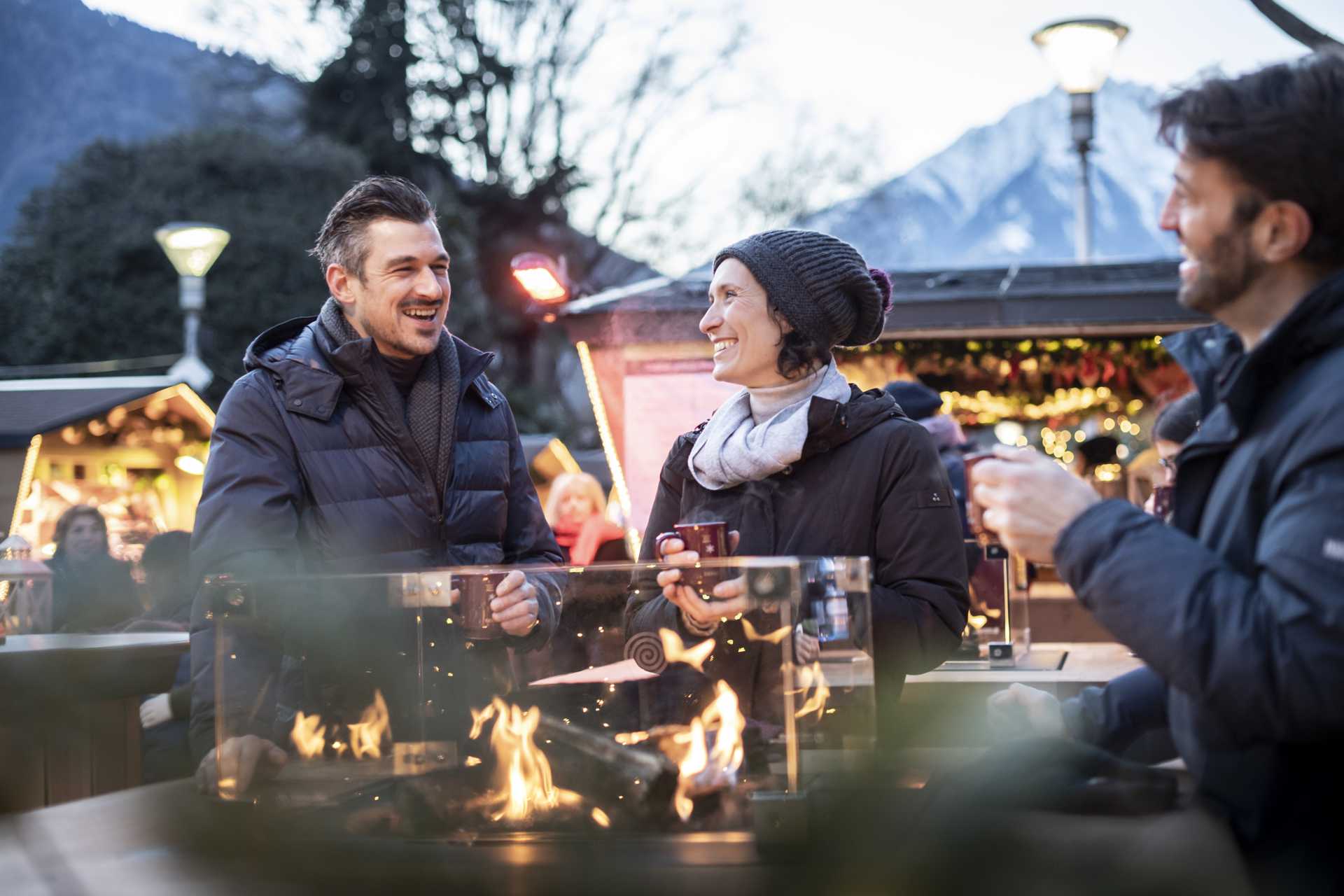 Three people drinking mulled wine and standing around a warming fire.